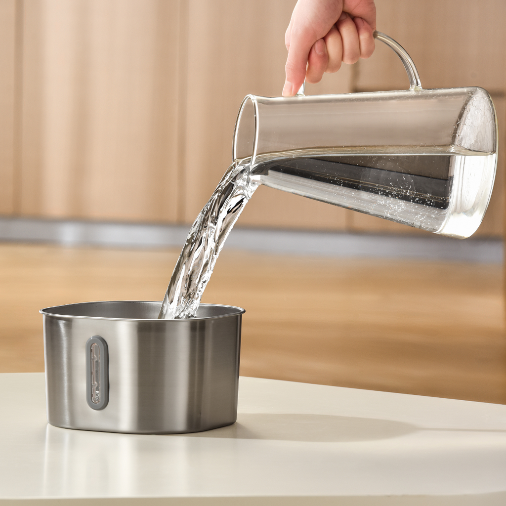 Clear glass pitcher being filled with water into a metal container on a kitchen counter.